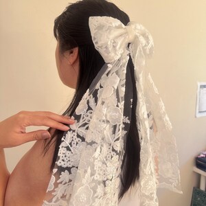 May include: White lace hair bow with a large bow and long tails. The bow is made of delicate lace with a floral pattern.