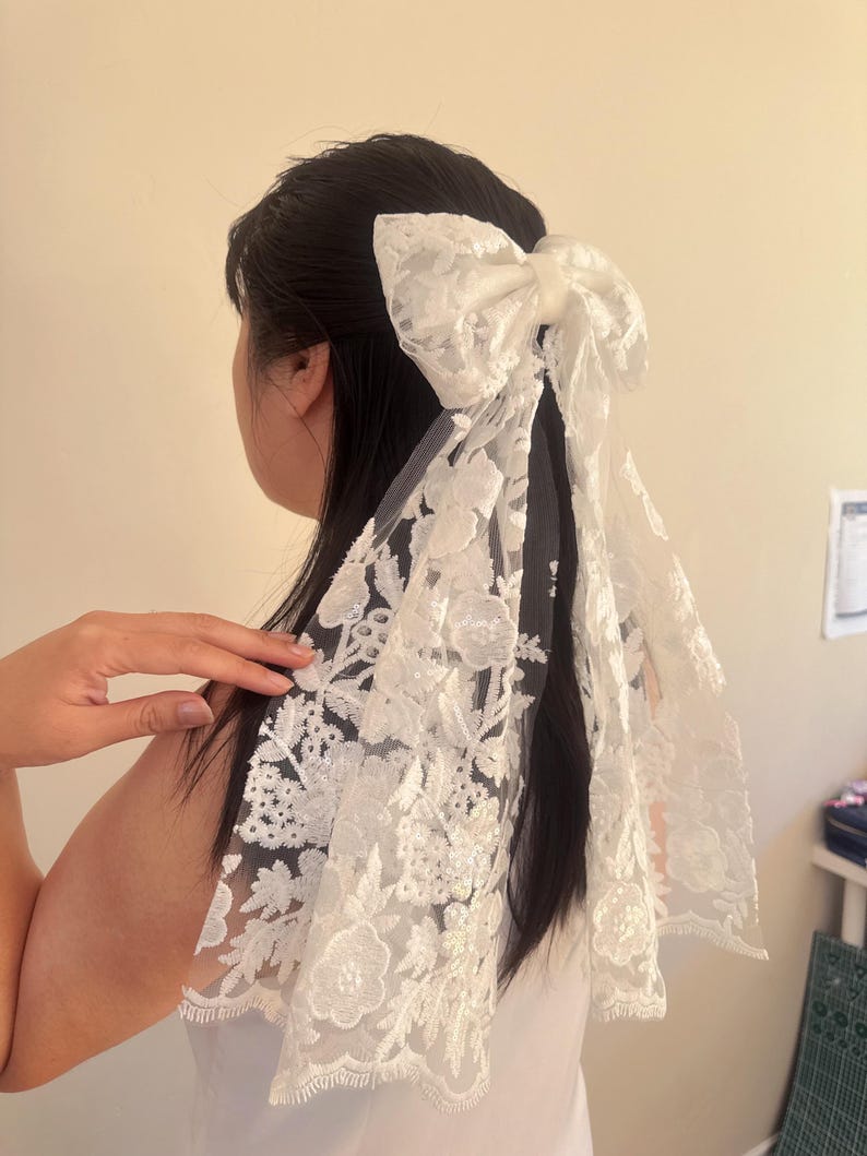 May include: White lace hair bow with a large bow and long tails. The bow is made of delicate lace with a floral pattern.