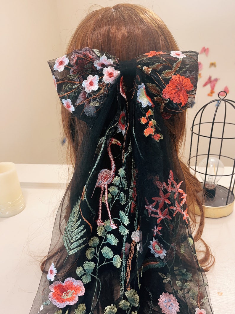 May include: Black sheer hair scarf with floral embroidery. The embroidery features pink, red, and green flowers, as well as a flamingo.