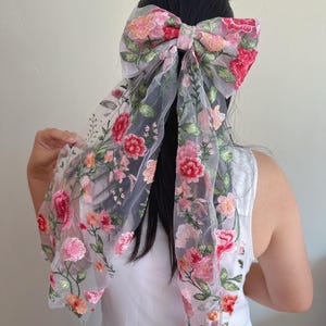 May include: A large, sheer hair bow with intricate floral embroidery in shades of pink, red, and peach. The bow is attached to a long, flowing fabric, creating a delicate and feminine accessory. The bow is worn in the hair.