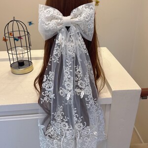 May include: A white lace bow hair accessory with a long, flowing veil. The bow is large and detailed, with intricate floral embroidery. The veil also features delicate floral patterns, creating an elegant and romantic look.