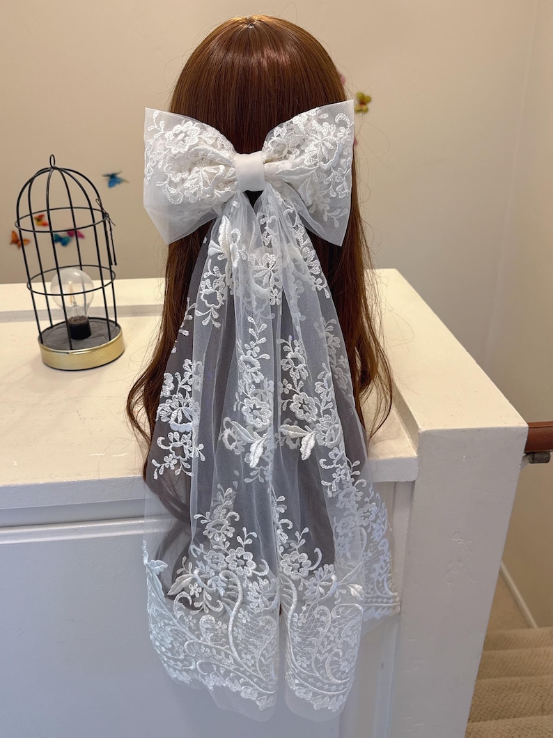May include: A white lace bow hair accessory with a long, flowing veil. The bow is large and detailed, with intricate floral embroidery. The veil also features delicate floral patterns, creating an elegant and romantic look.