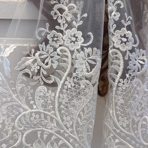 May include: Close-up of white lace fabric with intricate floral embroidery. The delicate design features detailed flowers and leaves, creating a textured pattern. The lace is likely used for bridal wear or other formal garments.