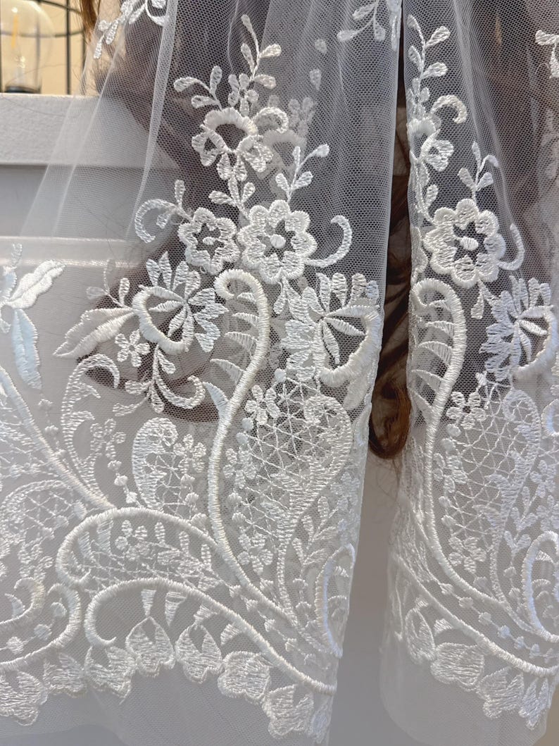 May include: Close-up of white lace fabric with intricate floral embroidery. The delicate design features detailed flowers and leaves, creating a textured pattern. The lace is likely used for bridal wear or other formal garments.