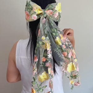 May include: A large, sheer hair bow with floral embroidery in yellow, pink, and orange. The bow is attached to a long, flowing fabric with the same floral design. The bow is worn in the hair.