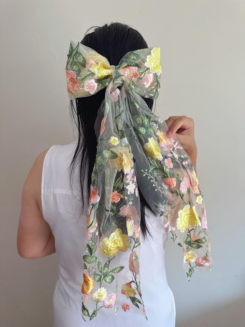 May include: A large, sheer hair bow with floral embroidery in yellow, pink, and orange. The bow is attached to a long, flowing fabric with the same floral design. The bow is worn in the hair.