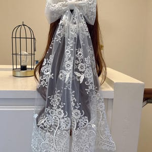 May include: A white lace hair bow with a large bow and flowing veil. The bow and veil feature intricate floral embroidery. The hair accessory is designed to be worn in the hair. The overall style is elegant and suitable for special occasions.