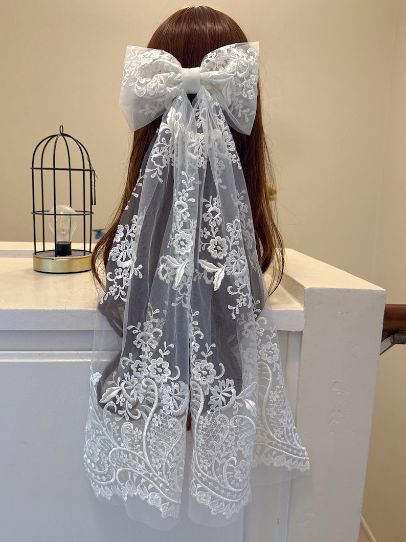 May include: A white lace hair bow with a large bow and flowing veil. The bow and veil feature intricate floral embroidery. The hair accessory is designed to be worn in the hair. The overall style is elegant and suitable for special occasions.