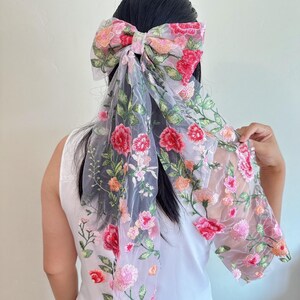 May include: A floral hair bow made of sheer, light grey fabric. The bow is embroidered with red, pink, and orange flowers and green leaves. The bow is tied in the hair, with the fabric ends flowing down the back.