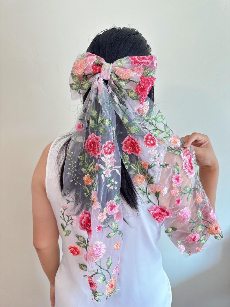 May include: A floral hair bow made of sheer, light grey fabric. The bow is embroidered with red, pink, and orange flowers and green leaves. The bow is tied in the hair, with the fabric ends flowing down the back.