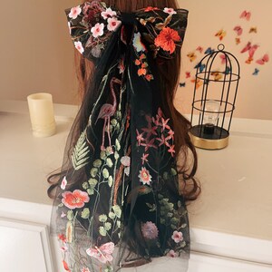 May include: Black sheer hair scarf with floral embroidery. The scarf features a large bow with pink, red, and blue flowers. The scarf also features a flamingo, butterflies, and other floral designs.