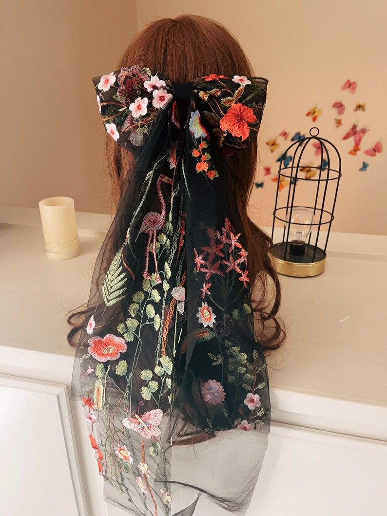 May include: Black sheer hair scarf with floral embroidery. The scarf features a large bow with pink, red, and blue flowers. The scarf also features a flamingo, butterflies, and other floral designs.