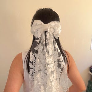 May include: White lace bridal hair bow with a large bow and a long flowing veil. The bow is tied in a knot at the back of the head.