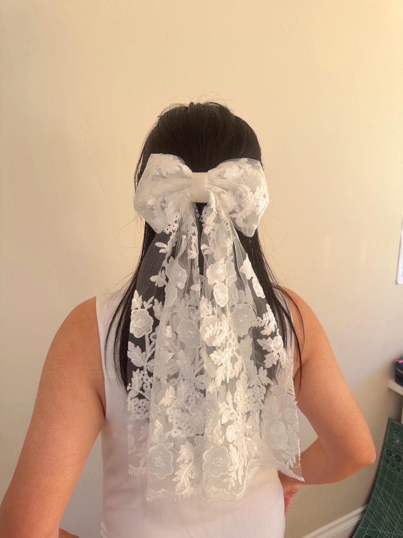 May include: White lace bridal hair bow with a large bow and a long flowing veil. The bow is tied in a knot at the back of the head.