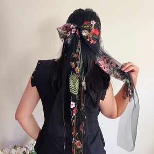 May include: Black sheer hair scarf with floral embroidery. The scarf is tied in a bow at the top of the head and the ends hang down the back.