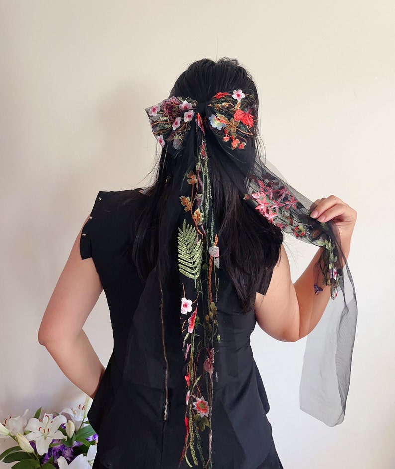 May include: Black sheer hair scarf with floral embroidery. The scarf is tied in a bow at the top of the head and the ends hang down the back.