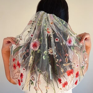 May include: A sheer white veil with colorful floral embroidery. The veil features red, pink, and green flowers with gold and brown accents. The veil is held up by a hair accessory.
