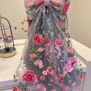 May include: A large, sheer hair bow with intricate floral embroidery in shades of pink, red, and orange. The bow is attached to a long, flowing veil, also adorned with embroidered flowers and green leaves. The design is delicate and feminine.