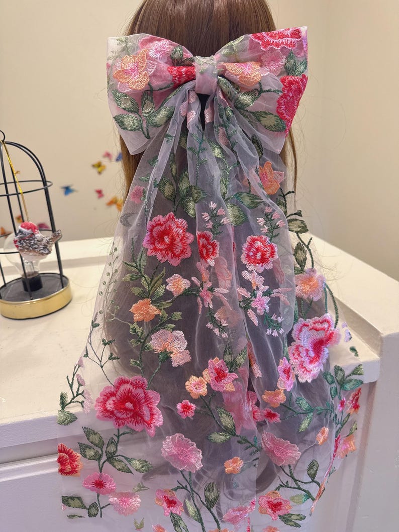 May include: A large, sheer hair bow with intricate floral embroidery in shades of pink, red, and orange. The bow is attached to a long, flowing veil, also adorned with embroidered flowers and green leaves. The design is delicate and feminine.