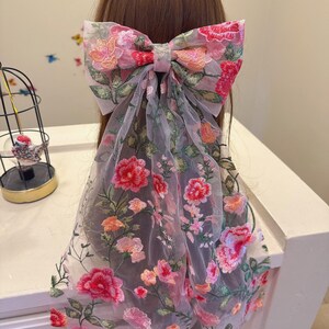 May include: A large hair bow with floral embroidery in pink, red, and orange hues. The bow is attached to a sheer fabric that drapes downwards. The design features detailed floral patterns, creating a delicate and feminine accessory.