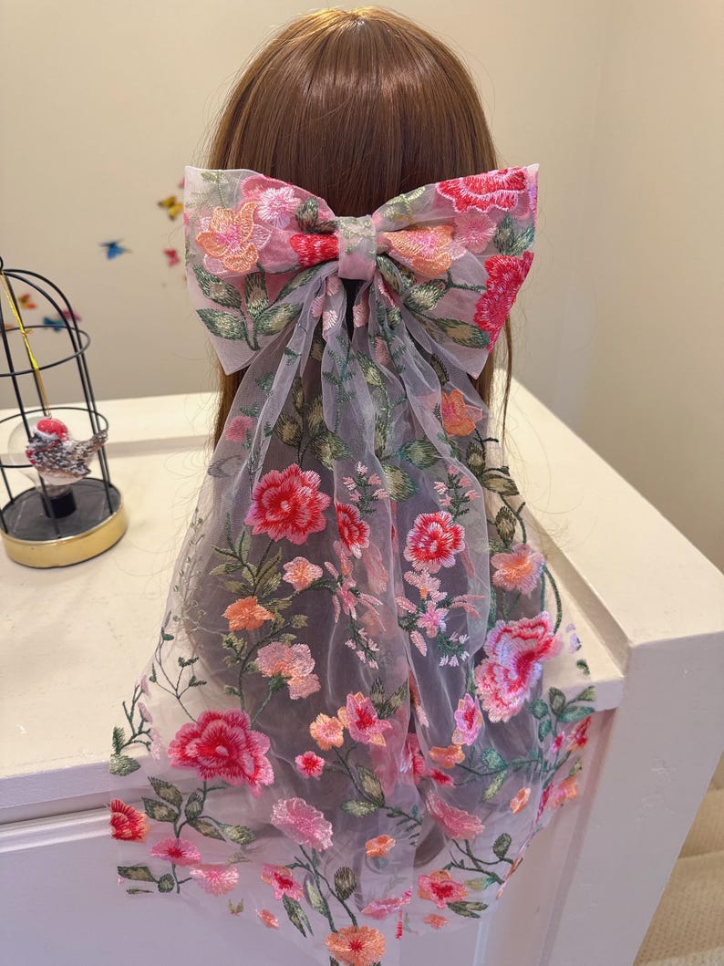 May include: A large hair bow with floral embroidery in pink, red, and orange hues. The bow is attached to a sheer fabric that drapes downwards. The design features detailed floral patterns, creating a delicate and feminine accessory.