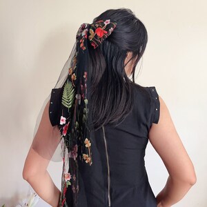 May include: A black dress with a floral embroidered sheer scarf tied in a bow at the back of the hair.