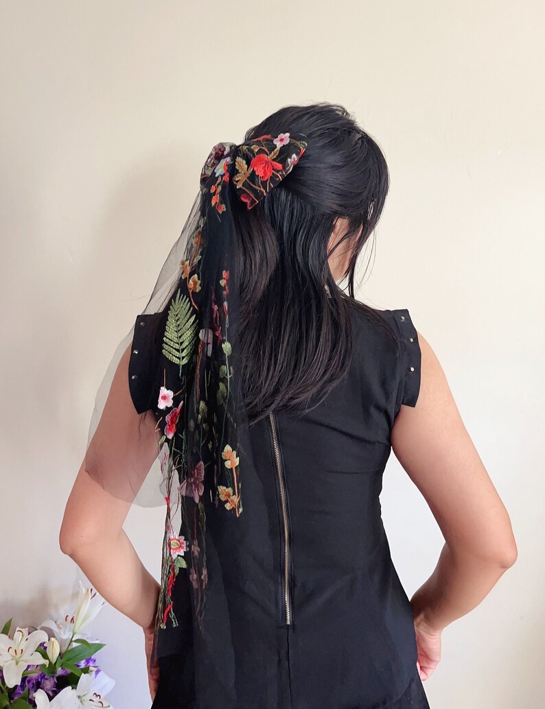 May include: A black dress with a floral embroidered sheer scarf tied in a bow at the back of the hair.