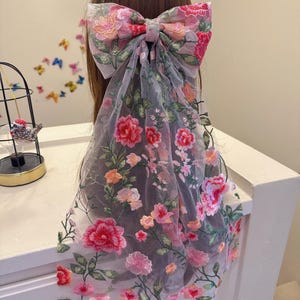 May include: A large, floral-embroidered bow hair accessory. The bow features a pink and red flower design with green leaves on a sheer, grey fabric. The bow is tied in the centre and drapes down the back.