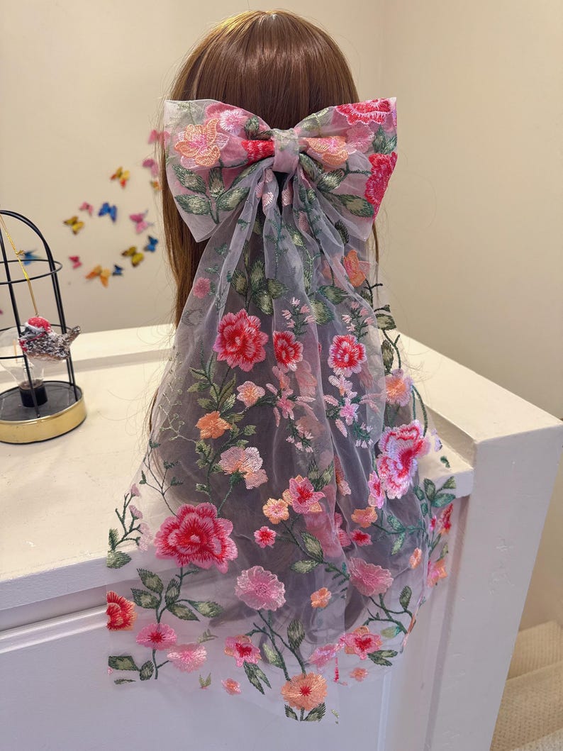 May include: A large, floral-embroidered bow hair accessory. The bow features a pink and red flower design with green leaves on a sheer, grey fabric. The bow is tied in the centre and drapes down the back.