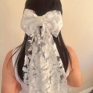 May include: A white lace bow hair clip with a large bow and a long, flowing tail. The bow is made of delicate lace with a floral pattern.