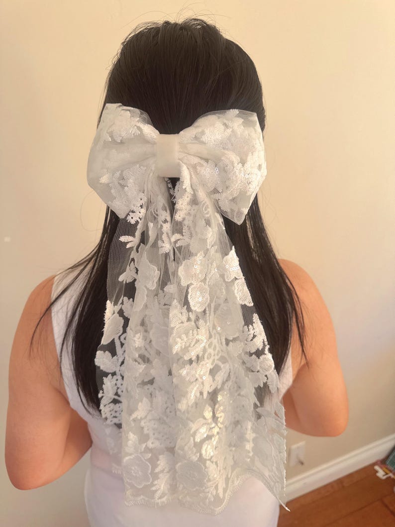 May include: A white lace bow hair clip with a large bow and a long, flowing tail. The bow is made of delicate lace with a floral pattern.