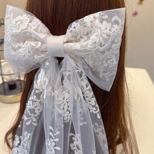 May include: A large white lace bow hair accessory. The bow features intricate floral embroidery and long, flowing tulle tails. The bow is secured with a satin band, creating a delicate and elegant look. The accessory is designed to be worn in the hair.