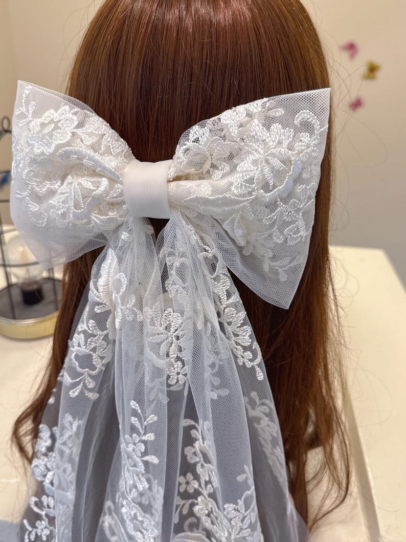 May include: A large white lace bow hair accessory. The bow features intricate floral embroidery and long, flowing tulle tails. The bow is secured with a satin band, creating a delicate and elegant look. The accessory is designed to be worn in the hair.