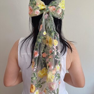 May include: A large, floral embroidered hair bow in shades of yellow, pink, and green. The bow is attached to a sheer, flowing fabric that drapes down. The design features detailed floral embroidery, creating a delicate and feminine accessory.