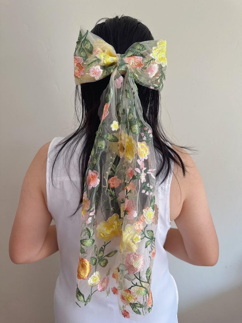 May include: A large, floral embroidered hair bow in shades of yellow, pink, and green. The bow is attached to a sheer, flowing fabric that drapes down. The design features detailed floral embroidery, creating a delicate and feminine accessory.
