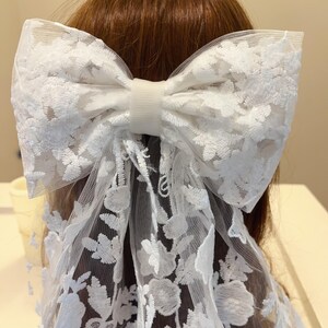 May include: A white lace bow hair clip with a floral pattern. The bow is tied with a white ribbon and has a white elastic band.