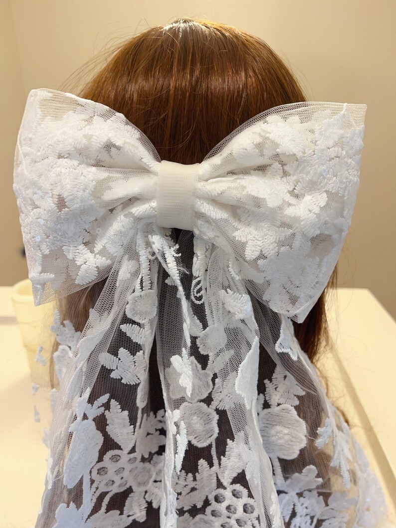May include: A white lace bow hair clip with a floral pattern. The bow is tied with a white ribbon and has a white elastic band.
