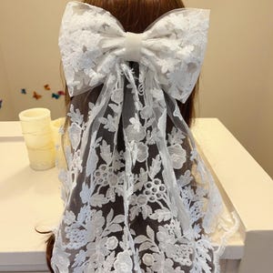 May include: White lace hair bow with a large bow and a long, flowing veil. The veil is decorated with a floral pattern.