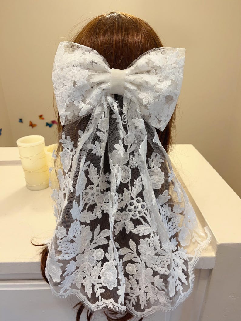 May include: White lace hair bow with a large bow and a long, flowing veil. The veil is decorated with a floral pattern.