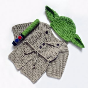 Könnte beinhalten: Ein gehäkeltes Baby-Outfit in Beige mit einer passenden grünen Häkelmütze. Die Mütze hat große Ohren und ähnelt einer beliebten fiktiven Figur. Ein kleines gehäkeltes Lichtschwert ist enthalten.