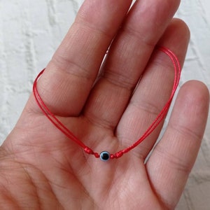 May include: A red string bracelet with a blue evil eye charm. The bracelet is tied around a person's wrist.