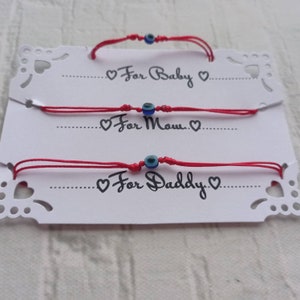 Può includere: Tre braccialetti di filo rosso con ciondoli blu a forma di occhio turco. I braccialetti sono etichettati "Per Baby", "Per Mamma" e "Per Papà".