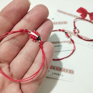 May include: Red string bracelet with a silver ladybug charm. The bracelet is tied with a knot and has a small red bead at the knot. The ladybug charm has black spots and white wings.