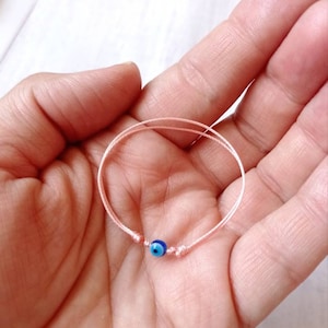 Puede incluir: Una pulsera de cuerda rosa con un dije azul de ojo turco.