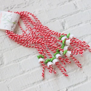 Op de afbeelding: Een bundel rood en wit touw met witte bloemvormige kralen en groene bladeren. Het touw is gebonden met een label waarop "Весела Великдень!" in het Cyrillisch staat.