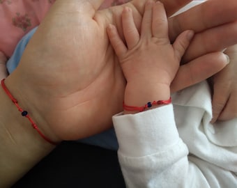 May include: A close-up of two wrists wearing red string bracelets with small blue beads. The bracelets are tied in a knot and are worn on the wrists of an adult and a baby.