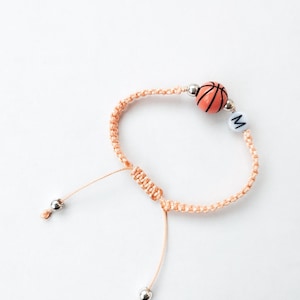 May include: A peach-colored friendship bracelet with a basketball charm and a silver bead with the letter "M" on it.
