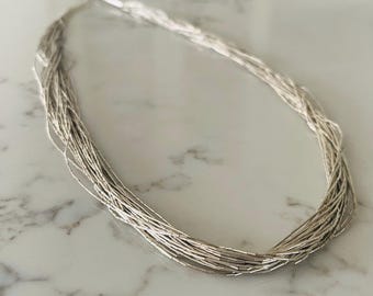 May include: A multi-strand liquid silver necklace composed of numerous thin, flexible chains featuring small, cylindrical metallic beads. The necklace has a bright, reflective silver finish and is arranged in a long, flowing, oval-shaped loop.
