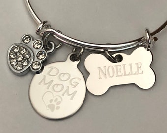 May include: A silver-toned charm bracelet with multiple charms. Charms include a paw print with clear crystals, a round charm with "Dog Mom" and a paw print, and a bone-shaped charm with the name "Noelle".