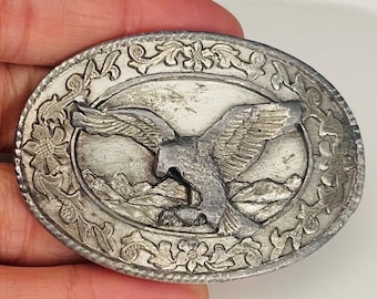 May include: A silver belt buckle with an eagle design. The eagle is in flight with its wings spread wide. The buckle is surrounded by a decorative border of flowers and leaves.
